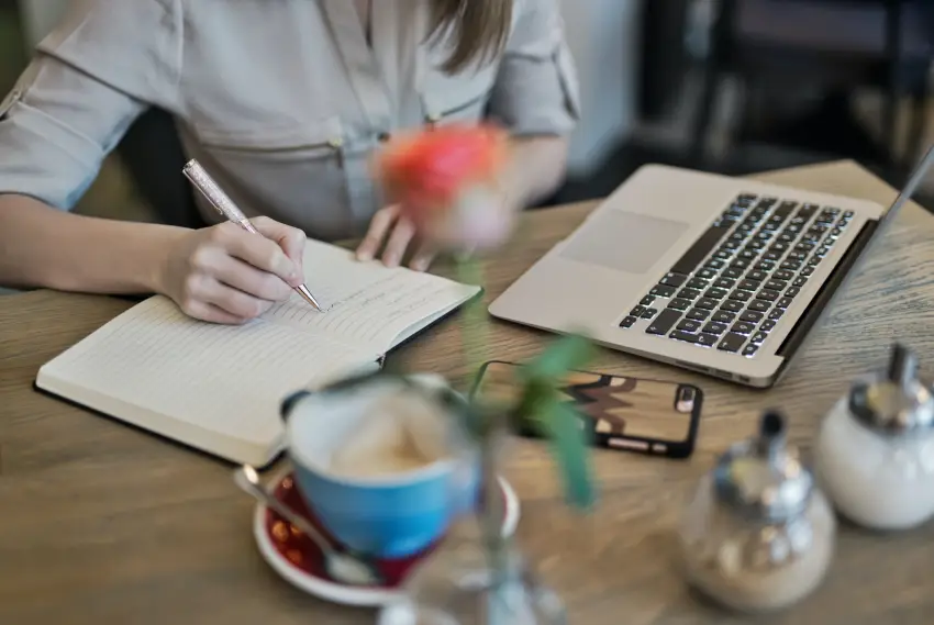Mujer redacta contenido para páginas web en una libreta con ordenador delante y taza de café